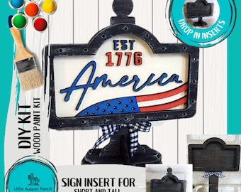 Puede incluir: Kit de bricolaje para letrero de madera con un letrero en blanco y negro que dice "Est 1776 America" con una bandera estadounidense roja, blanca y azul. El letrero está en un soporte negro con una cinta a cuadros en blanco y negro. El kit incluye pintura y un pincel.