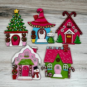 May include: Five wooden Christmas ornaments shaped like houses. Each house is painted in different colors, including pink, green, blue, and red. The houses have candy cane details and are decorated with glitter. One house has the words "North Pole" written on it.