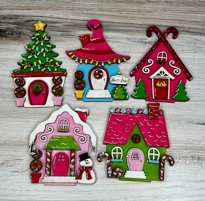 May include: Five wooden Christmas ornaments shaped like houses. Each house is painted in different colors, including pink, green, blue, and red. The houses have candy cane details and are decorated with glitter. One house has the words "North Pole" written on it.