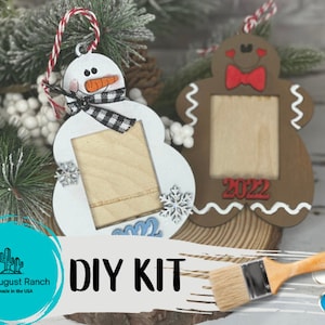 May include: DIY Christmas ornament kit featuring a snowman and gingerbread man with picture frames. The kit includes paint, brushes, and the wooden ornaments. The ornaments are ready to be painted and personalized.