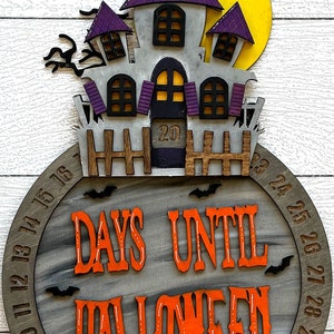 May include: A wooden Halloween countdown clock with a purple and gray haunted house design. The clock features a circular wooden base with the numbers 1-31 around the edge. The text "Days Until Halloween" is written in orange letters on the clock face.