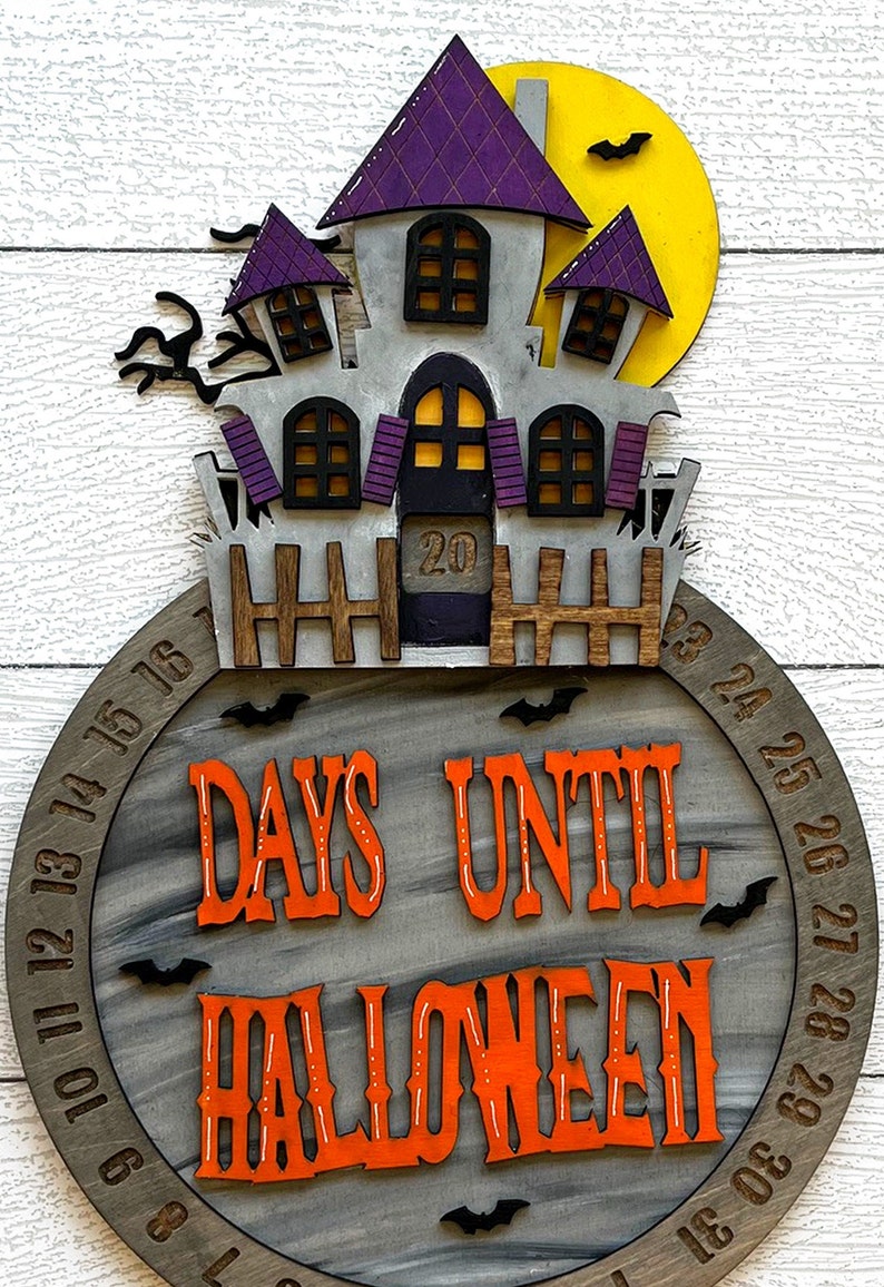 May include: A wooden Halloween countdown clock with a purple and gray haunted house design. The clock features a circular wooden base with the numbers 1-31 around the edge. The text "Days Until Halloween" is written in orange letters on the clock face.