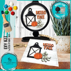May include: A DIY wood paint kit featuring a black lantern, orange pumpkin, and the words "HELLO FALL." The kit includes paint colors and a paintbrush. The design is on a white wooden surface. The kit is from Little August Ranch.