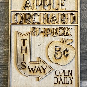 May include: A wooden sign with the text "APPLE ORCHARD U-PICK THIS WAY OPEN DAILY" and a red apple with a 5&cent; price tag.