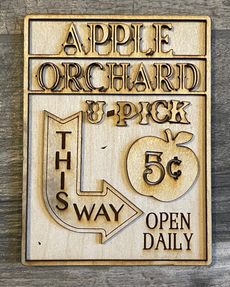 May include: A wooden sign with the text "APPLE ORCHARD U-PICK THIS WAY OPEN DAILY" and a red apple with a 5&cent; price tag.