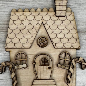 May include: A wooden cutout of a gingerbread house with candy cane accents. The house has a scalloped roof, a chimney, a door with a small window, and two windows on the sides.