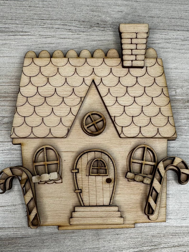 May include: A wooden cutout of a gingerbread house with candy cane accents. The house has a scalloped roof, a chimney, a door with a small window, and two windows on the sides.