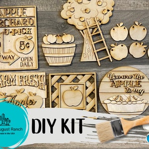 May include: DIY craft kit featuring wooden cutouts of an apple orchard theme. The kit includes signs that say "Apple Orchard", "U-Pick 5&cent;", "Farm Fresh", and "You are the Apple to my". There are also cutouts of an apple tree, ladder, apples, and an apple pie.