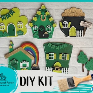 May include: DIY kit for creating five miniature houses with a leprechaun theme. The kit includes a pot of gold, a rainbow, and two trees. The houses are painted green and have shamrock details. The kit also includes paint and a paintbrush.