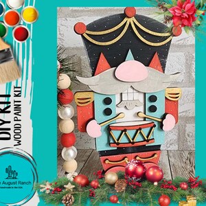 May include: DIY wood paint kit featuring a nutcracker design. The nutcracker is painted in shades of blue, red, and black with a gold trim. The kit includes a wooden nutcracker cutout, paint, and brushes. The nutcracker is holding a drum and has a mustache and a pink nose.