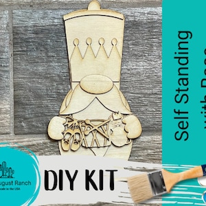 May include: DIY kit for a wooden gnome ornament. The gnome is holding a paintbrush and a paint palette. The gnome is wearing a crown and has a beard. The text "Self Standing with Base" and "DIY KIT" is visible in the image.