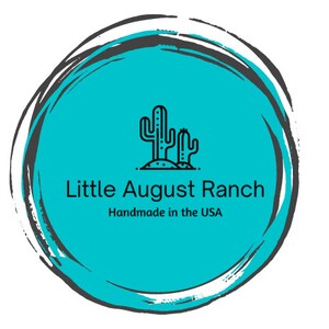 May include: A turquoise circle with a black outline and a white brushstroke effect. Inside the circle is a black line drawing of two cacti with the text "Little August Ranch" and "Handmade in the USA" below.