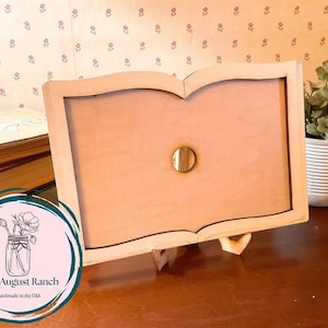 May include: A wooden book-shaped frame with a light brown interior and a small, round, gold-colored knob. The frame is propped up on a wooden stand. The Little August Ranch logo is in the lower left corner.