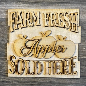 May include: A wooden sign with the text "Farm Fresh Apples Sold Here" in a rustic font. The sign is painted in a light brown color and features three stylized apples.