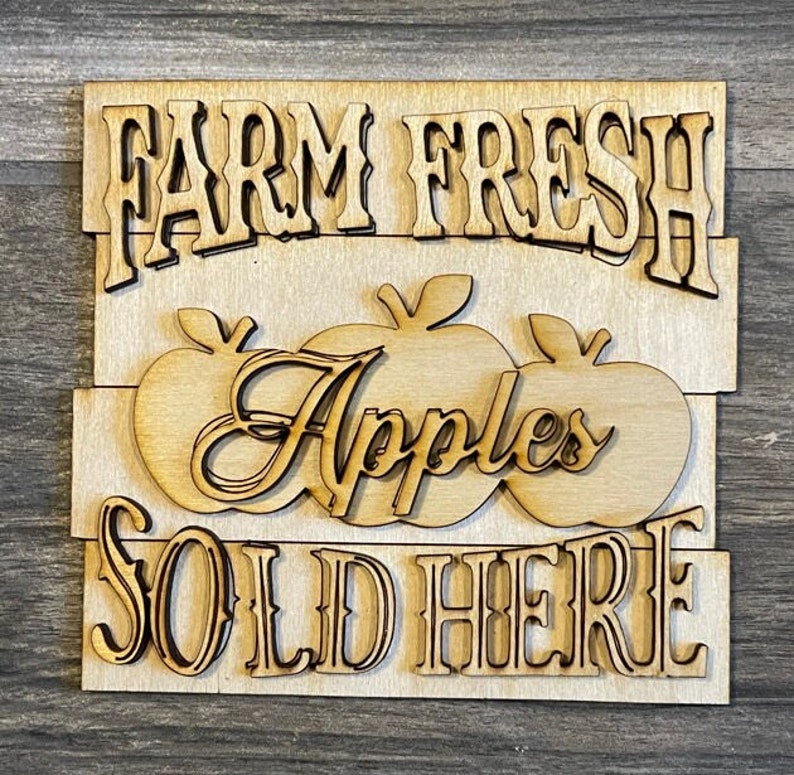 May include: A wooden sign with the text "Farm Fresh Apples Sold Here" in a rustic font. The sign is painted in a light brown color and features three stylized apples.