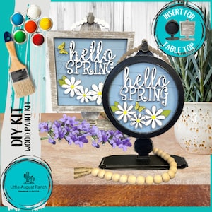 May include: DIY kit for a wooden sign with the words "hello spring" in white letters on a blue background. The sign is decorated with white daisies and butterflies. The kit includes paint, a wooden sign, and a wooden stand.