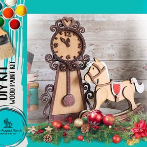 May include: DIY wood paint kit featuring a wooden clock and rocking horse. The clock has a brown face with white numbers and hands. The rocking horse is brown with a red saddle. The kit is surrounded by Christmas decorations, including a pine branch, red berries, and a poinsettia.