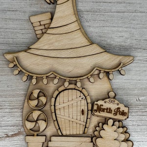 May include: Wooden cutout of a whimsical elf house with a pointed roof, a door, candy cane decorations, and a sign that reads "North Pole".