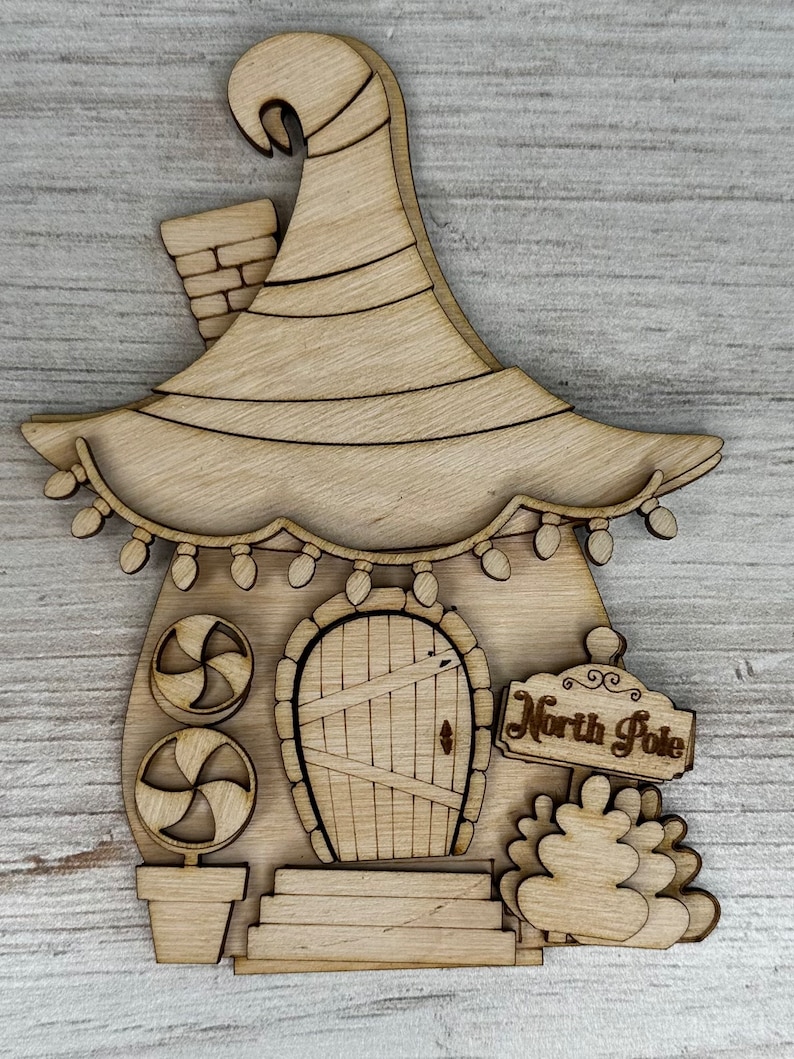May include: Wooden cutout of a whimsical elf house with a pointed roof, a door, candy cane decorations, and a sign that reads "North Pole".