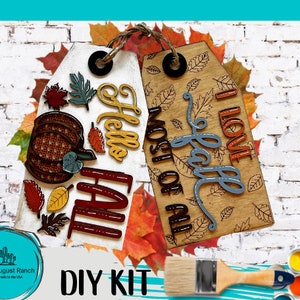 May include: DIY craft kit featuring two wooden tags with fall-themed designs. One tag says "Hello Fall" with a pumpkin and leaves, the other says "I love fall most of all". The kit includes paint and a paintbrush.