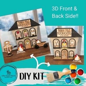 May include: A DIY kit for a 3D wooden nutcracker shop with a sign that reads "North Pole Nutcrackers & Nut Roasters". The kit includes paint, a paintbrush, and all the pieces needed to assemble the shop.