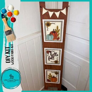 May include: A wooden DIY paint kit featuring three panels with fall-themed designs. The top panel has a banner with white triangles and a pumpkin with sunflowers. The middle panel says "hello fall" with a sunflower. The bottom panel has a pumpkin with sunflowers.