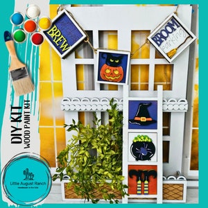 May include: DIY wood paint kit featuring a white wooden house with Halloween-themed decorations. Includes a banner with "Witches Brew" and "Broom Parking" signs, a pumpkin, a witch's hat, and a cauldron. Paint colors and a paintbrush are also included.