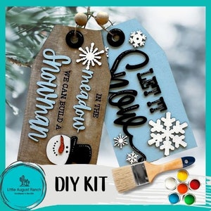 May include: DIY kit for creating two wooden signs with winter themes. The signs are painted blue and brown and feature the text "Let it Snow" and "In the Meadow We Can Build a Snowman". The kit includes paint, brushes, and stencils.