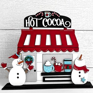 May include: Wooden cutout of a hot cocoa stand with a red roof and white trim. The sign reads "Hot Cocoa". There are two snowmen on either side of the stand, one holding a red cup and the other holding a red scarf.