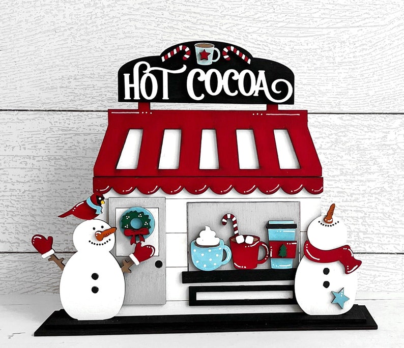 May include: Wooden cutout of a hot cocoa stand with a red roof and white trim. The sign reads "Hot Cocoa". There are two snowmen on either side of the stand, one holding a red cup and the other holding a red scarf.