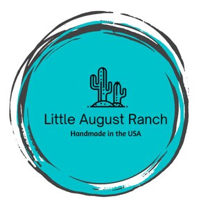 May include: A turquoise blue circle with a black outline and a white brushstroke effect. Inside the circle is a black line drawing of two cacti with the text "Little August Ranch" and "Handmade in the USA" below.