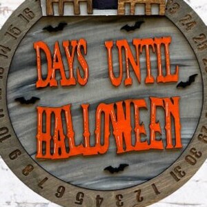 May include: A wooden countdown clock with a gray background and orange text that reads "Days Until Halloween". The clock has black bats and a black silhouette of a house on the top.