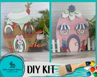 May include: DIY kit for creating two Easter egg-shaped houses. One house is brown with green accents and the other is pink with purple accents. Both houses have bunny windows and a carrot shop sign. The kit includes paint and a paintbrush.