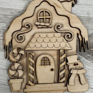 May include: A wooden cutout of a gingerbread house with a snowman standing next to it. The house has a candy cane plant in front of it and a snowy roof.