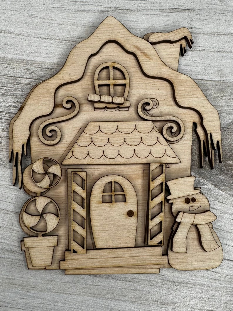 May include: A wooden cutout of a gingerbread house with a snowman standing next to it. The house has a candy cane plant in front of it and a snowy roof.