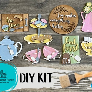 May include: DIY craft kit for making tea-themed wooden signs. The kit includes a teapot, teacups, a tray with tea time text, a sign with the text "Tea makes everything better", a sign with the text "but first tea", a sugar bowl, a tray with croissants, and a paintbrush with paint pots.
