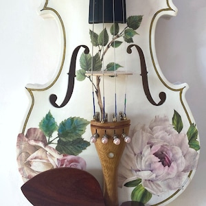 Pu&ograve; includere: Un violino bianco con un design floreale con rose rosa e foglie verdi. Il violino ha un manico in legno scuro e un poggiamento per il mento marrone.
