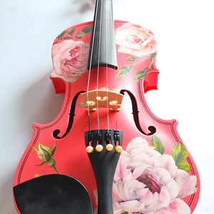 Pu&ograve; includere: Un violino rosso con un design floreale con rose rosa e foglie. Il violino ha un manico nero e un poggiamento per il mento nero.