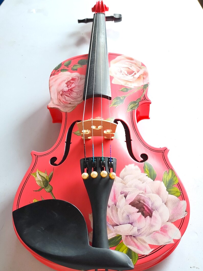 Pu&ograve; includere: Un violino rosso con un design floreale con rose rosa e foglie. Il violino ha un manico nero e un poggiamento per il mento nero.