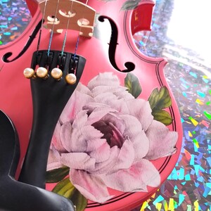 Pu&ograve; includere: Un violino rosa con un disegno floreale che presenta una grande peonia bianca. Il violino ha un manico nero e un set di bottoni bianchi perlati.