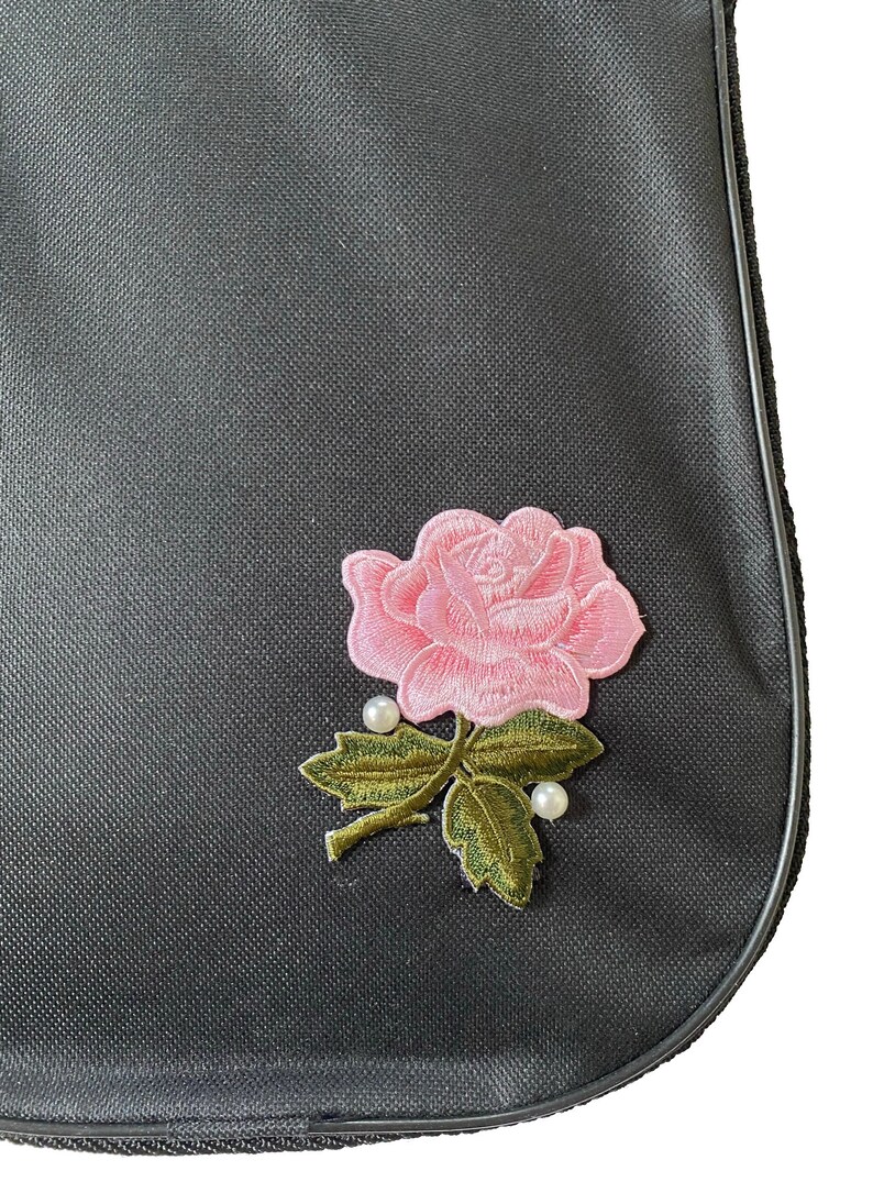 Pu&ograve; includere: Una borsa in tessuto nero con una rosa ricamata rosa e dettagli di perle.
