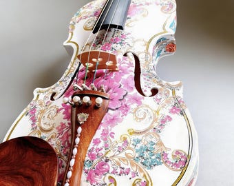 May include: A white violin with a floral design featuring pink flowers and gold accents. The instrument has a wooden bridge, fingerboard, and chin rest. The design includes ornate scrollwork and a decorative tailpiece.