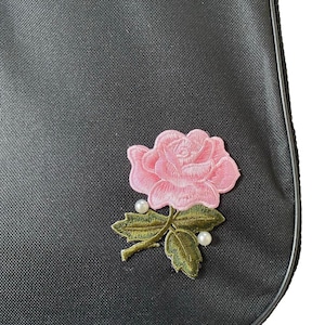 A close-up view of a black fabric with a pink rose embroidered on it, featuring a green leaf and pearl accents.