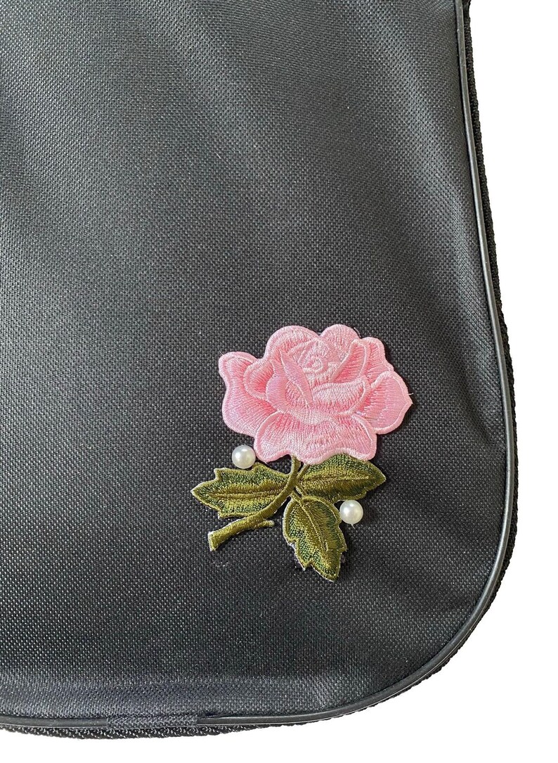 A close-up view of a black fabric with a pink rose embroidered on it, featuring a green leaf and pearl accents.