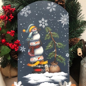 May include: A whimsical, hand-painted wooden sign featuring a snowman balancing on skis, with a small Christmas tree. The background is a dark blue with white snowflakes. The sign includes the text "Marika Moretti 2023".