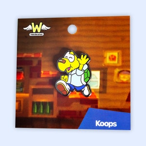 Peut inclure: Une épingle en émail jaune et bleue représentant un personnage de tortue de dessin animé portant une chemise blanche et une carapace verte. L'épingle est sur un fond brun avec le texte "Wonderate!" et "Koops" en blanc.