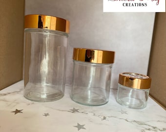 May include: Three glass jars with gold lids. The jars are different sizes and are arranged from largest to smallest. The jars are on a white marble surface.