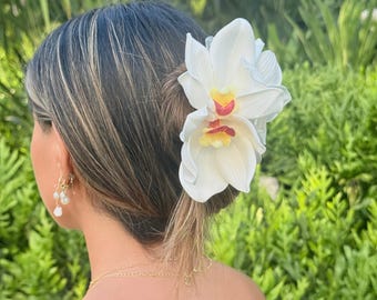 Könnte beinhalten: Eine Haarspange mit einer großen, weißen Orchideenblüte mit gelben und roten Zentren. Die Blütenblätter haben eine glatte, realistische Textur und die Blume ist an einem dunkel gefärbten Haarclip-Mechanismus befestigt, der das Haar hält.