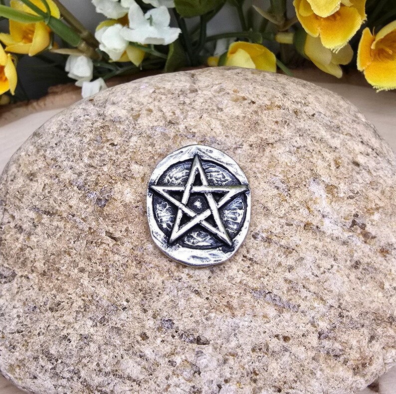 Puede incluir: Un amuleto de pentagrama plateado con un dise&ntilde;o de estrella de cinco puntas. El amuleto est&aacute; colocado sobre una piedra texturizada de color marr&oacute;n claro, con flores amarillas y blancas en el fondo. El amuleto tiene un borde circular.
