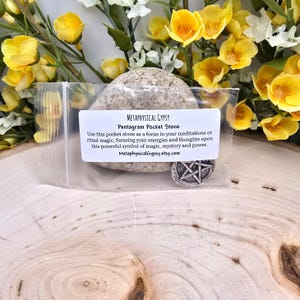 Puede incluir: Una peque&ntilde;a piedra gris lisa en una bolsa de pl&aacute;stico transparente con una etiqueta que dice "METAPHYSICAL GYPSY Pentagram Pocket Stone". La piedra se exhibe sobre una superficie de madera, rodeada de flores amarillas y blancas. Tambi&eacute;n es visible un peque&ntilde;o amuleto de pentagrama plateado.